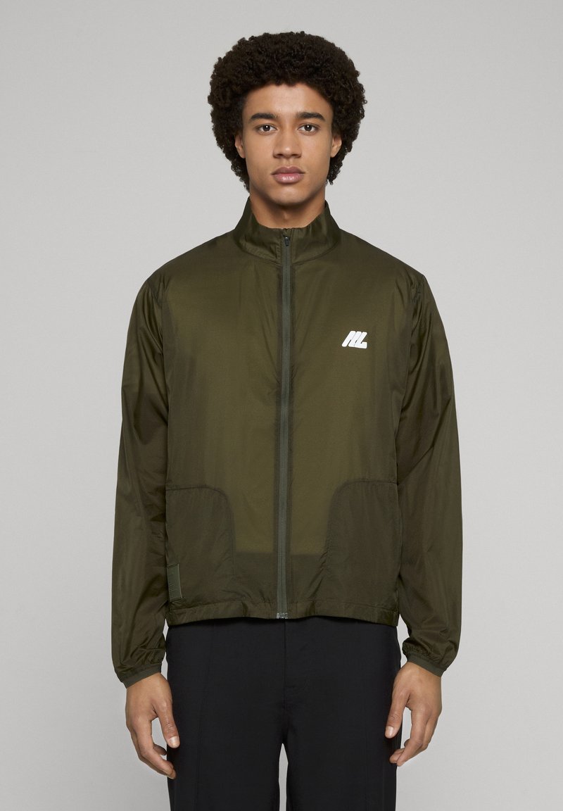 Lekka kurtka w kolorze oliwkowej zieleni z wysokim kołnierzem, zapięciem na suwak, bocznymi kieszeniami i białym logo na piersi. Gładka, nylonowa faktura.
