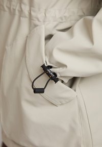 Beige waterdichte jas met een gestructureerde stof, elastische zaksluiting met zwarte toggle en koord, en zichtbare stikseldetails.