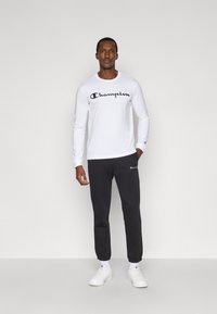 Maglietta bianca a maniche lunghe con logo "Champion" nero, abbinata a pantaloni della tuta grigio scuro e scarpe da ginnastica bianche. Design semplice e confortevole.