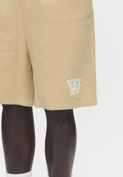 Shorts beiges à texture gaufrée avec un logo "W" brodé blanc sur la jambe gauche. Design simple, coupe jusqu'aux genoux, matériel léger.