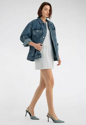 Frau in einem weißen Kleid, übergroßer blauer Jeansjacke und grauen Slingback-High Heels, die mit einer Hand in der Jackentasche geht.