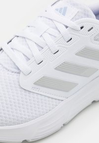 Zapatilla deportiva blanca con parte superior de tejido texturizado, cordones blancos y tres rayas plateadas en el costado cerca de la suela.