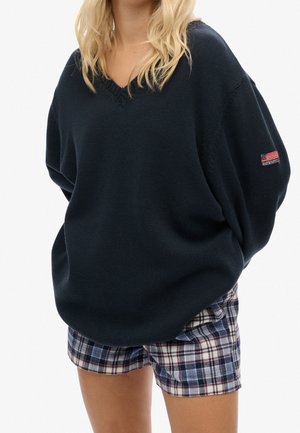Kobieta o blond włosach ubrana w luźny, ciemnoniebieski sweter z dekoltem w serek i niebieskie, białe oraz czerwone kraciaste szorty, trzymająca ręce za plecami.