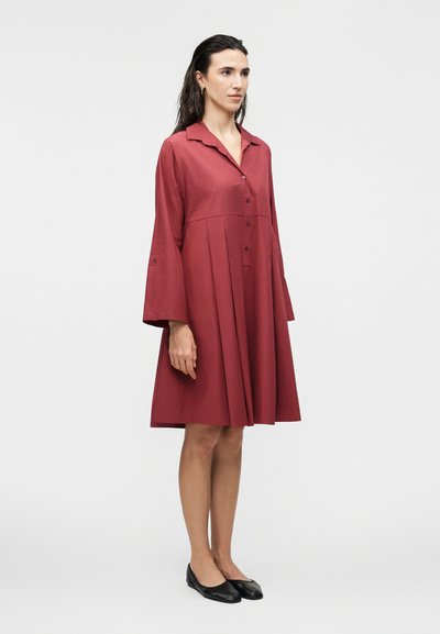 Γυναίκα που στέκεται πλευρικά φορώντας ένα άνετο κόκκινο φόρεμα με κουμπιά, φαρδιά μακριά μανίκια και μαύρα επίπεδα παπούτσια, σε απλό λευκό φόντο.