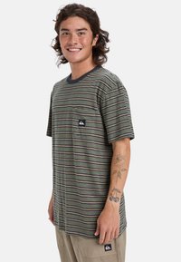 Kortærmet stribet t-shirt i grøn, marineblå og creme, med en lomme på venstre side og et logo-patch. Blødt bomuldsmateriale.