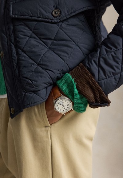 Main dans la poche d’un pantalon beige portant une montre en cuir marron, un pull en maille verte, un poignet en velours côtelé marron et une veste matelassée bleu foncé.