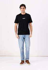 Sort bomulds T-shirt med korte ærmer og rund hals, med hvid tekst henover brystet. Båret med lyseblå jeans og brune sneakers.