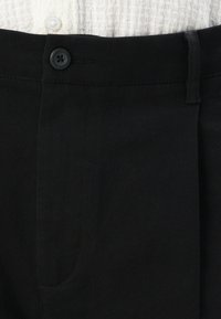 Schwarze Hose mit glatter Textur, ausgestattet mit einem Knopfverschluss und einer Fronttasche. Der Bund ist für Langlebigkeit verstärkt.
