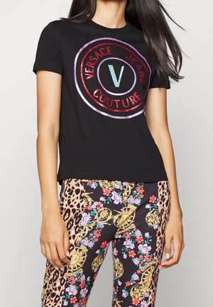 Femme portant un t-shirt noir Versace Jeans Couture avec un logo pailleté et un pantalon coloré à imprimé floral et léopard, debout devant un fond uni.