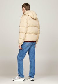 Beige puffer jacket with a hood, quilted design, elastic cuffs, worn over blue jeans and grey sneakers. The jacket has a relaxed fit.
