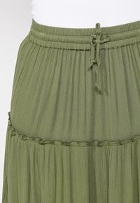 Jupe plissée vert olive avec taille élastique et lien coulissant, présentant un détail subtil de couture froncée sous la taille.