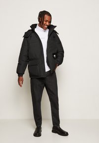 Giacca a vento nera con zip frontale, cappuccio regolabile con coulisse e due tasche anteriori; indossata sopra un dolcevita bianco e jeans scuri.