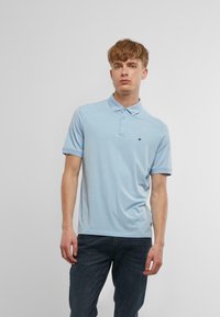 Gaiši zils polo krekls ar īsām piedurknēm, apkakli un trīs pogām pie priekšpuses. Uz kreisā krūts ir neliels melns logotips. Gluda tekstūra.