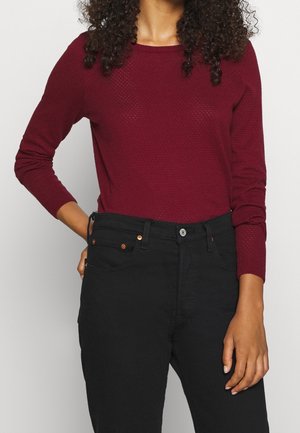 Femme portant un haut rouge foncé texturé à manches longues et un jean noir taille haute, se tenant avec une main sur la hanche et l'autre détendue.