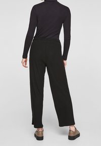 Personne portant un pantalon large côtelé noir, un haut noir à manches longues et des baskets à plateforme marron, debout devant un fond gris uni.