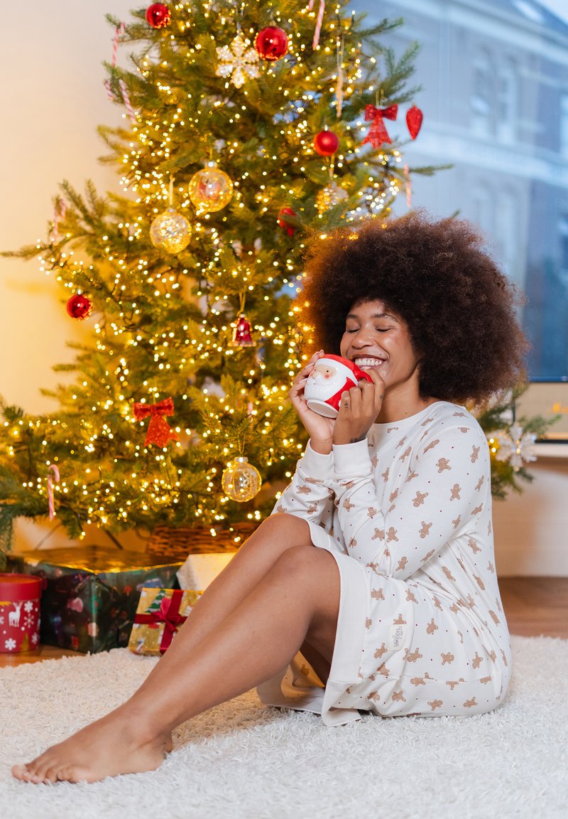 Witte nachtjapon met print van een peperkoekmannetje, zachte stof, tot op de knie, gedragen door een persoon die een kerstbeker vasthoudt, zittend op een zacht wit vloerkleed, kerstboom op de achtergrond.