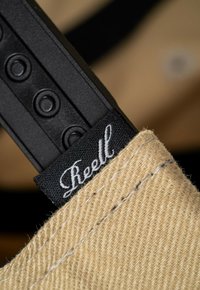 Canvas-Tasche mit beiger Textur, ausgestattet mit einem schwarzen Riemen mit runden Beschlägen und einem schwarzen Etikett mit weißer Schriftzugmarkierung.