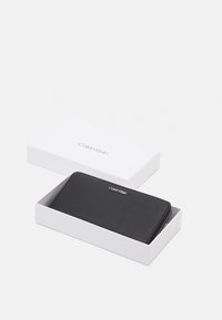 Portefeuille en cuir noir avec une finition texturée, fermeture éclair et logo Calvin Klein. Présenté dans une boîte blanche.