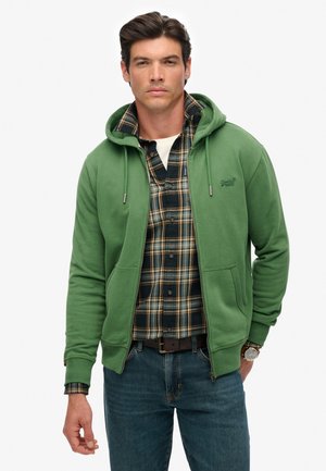 Man in een groene zip-up hoodie over een donkergroen en geel geruit shirt met blauwe jeans en een bruine horloge, staande tegen een witte achtergrond.