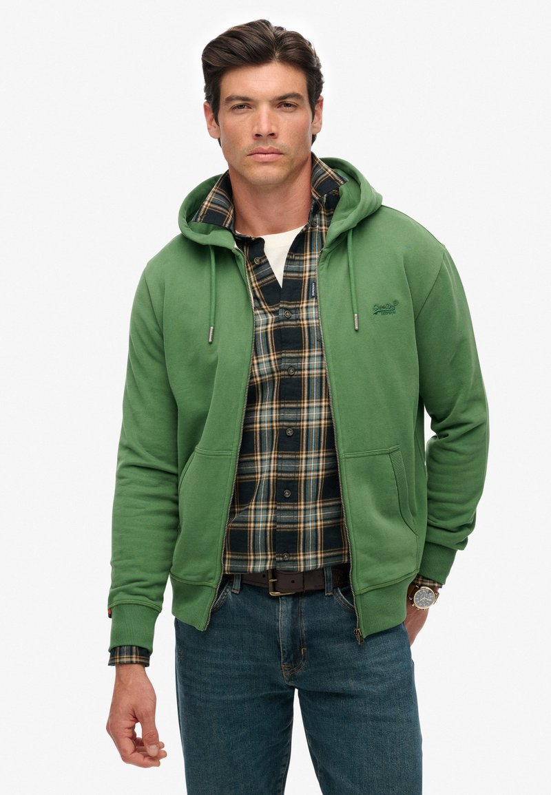 Hombre vistiendo una sudadera verde con cremallera sobre una camisa de cuadros en tonos verde oscuro y amarillo, con jeans azules y un reloj marrón, de pie contra un fondo blanco.