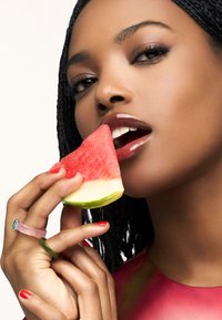 Modelka trzyma kawałek arbuza, ma na ustach błyszczącą bordową pomadkę i czerwony lakier na paznokciach oraz różowo-zielony pierścionek na palcach.