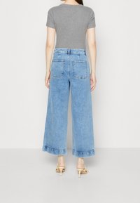 Nainen, jolla on yllä vaaleansiniset leveälahkeiset farkut, harmaa tiukka lyhytnpulloveri ja beige korkokengät, seisoo selin tasaista taustaa vasten.