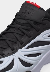 Μαύρο αθλητικό παπούτσι με περίπλοκο λευκό κυματιστό σχέδιο στη σόλα, μαύρα κορδόνια, κόκκινη ετικέτα στη γλώσσα και ανάγλυφο λογότυπο στη μαλακή γλώσσα.