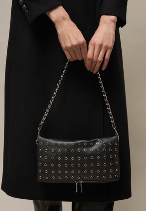 Ruky držiace čiernu koženú kabelku so striebornými očkami a retiazkovým remienkom na pozadí čierneho kabáta.