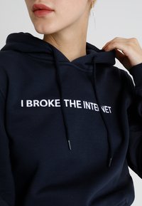 Tamna plava dukserica od mješavine pamuka, s kengur džepom i vezicama, s bijelim natpisom "POKVARIO SAM INTERNET" istaknutim.