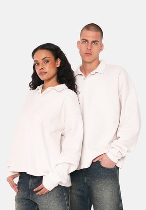 Light pink long-sleeve polo shirts with a soft texture, featuring buttoned collar and subtle embroidered details. Worn with dark jeans.