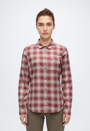 Une femme porte une chemise boutonnée à carreaux rouges et gris avec une poche poitrine, ainsi qu’un pantalon marron, sur un fond uni et clair.