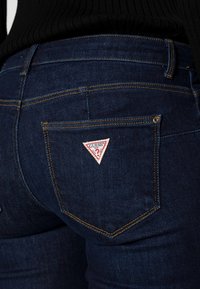 Jeans de mezclilla azul con una vista en primer plano del bolsillo trasero que presenta un parche triangular del logo "Guess" en rojo y blanco, y costuras amarillas.