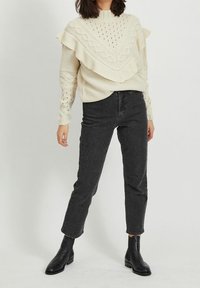 Pull épais crème avec un motif torsadé et un détail à volants superposés, associé à un jean taille haute gris foncé et des bottines noires.