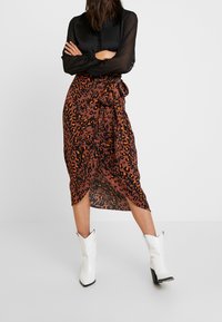 Femme les bras croisés portant une blouse noire transparente, une jupe enveloppante à motifs léopard brun et orange, et des bottes à talons blancs, debout.