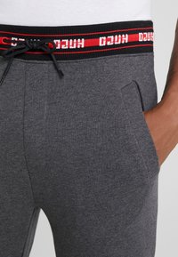 Graue Jogginghose mit einem elastischen Bund in Schwarz und Rot. Zu den Eigenschaften gehören Seitentaschen und ein Kordelzug zur Anpassung. Geschmeidige Textur.