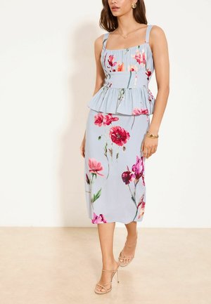 Kobieta w jasnoniebieskiej sukience midi bez rękawów z różowymi i czerwonymi kwiatowymi wzorami oraz beżowych sandałach na wysokim obcasie, stojąca przy białej ścianie.
