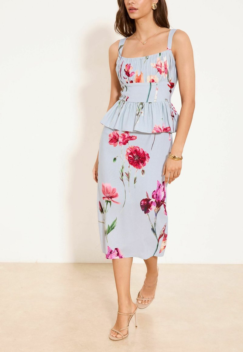 Frau trägt ein hellblaues ärmelloses Midikleid mit rosa und roten Blumenmustern und beigen Sandalen mit hohen Absätzen, steht an einer weißen Wand.