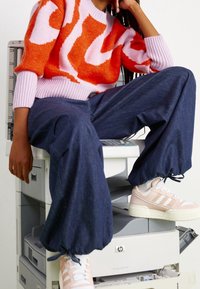 Pull en tricot à motif abstrait orange et rose, assorti à un pantalon en denim ample et des baskets rose clair avec des rayures blanches, assis sur une imprimante.