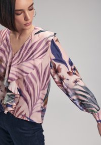 Blouse à motif floral en rose pâle, ornée de volants le long de l'encolure et des manches, avec des accents bleus et bruns. Fabriquée en tissu léger.