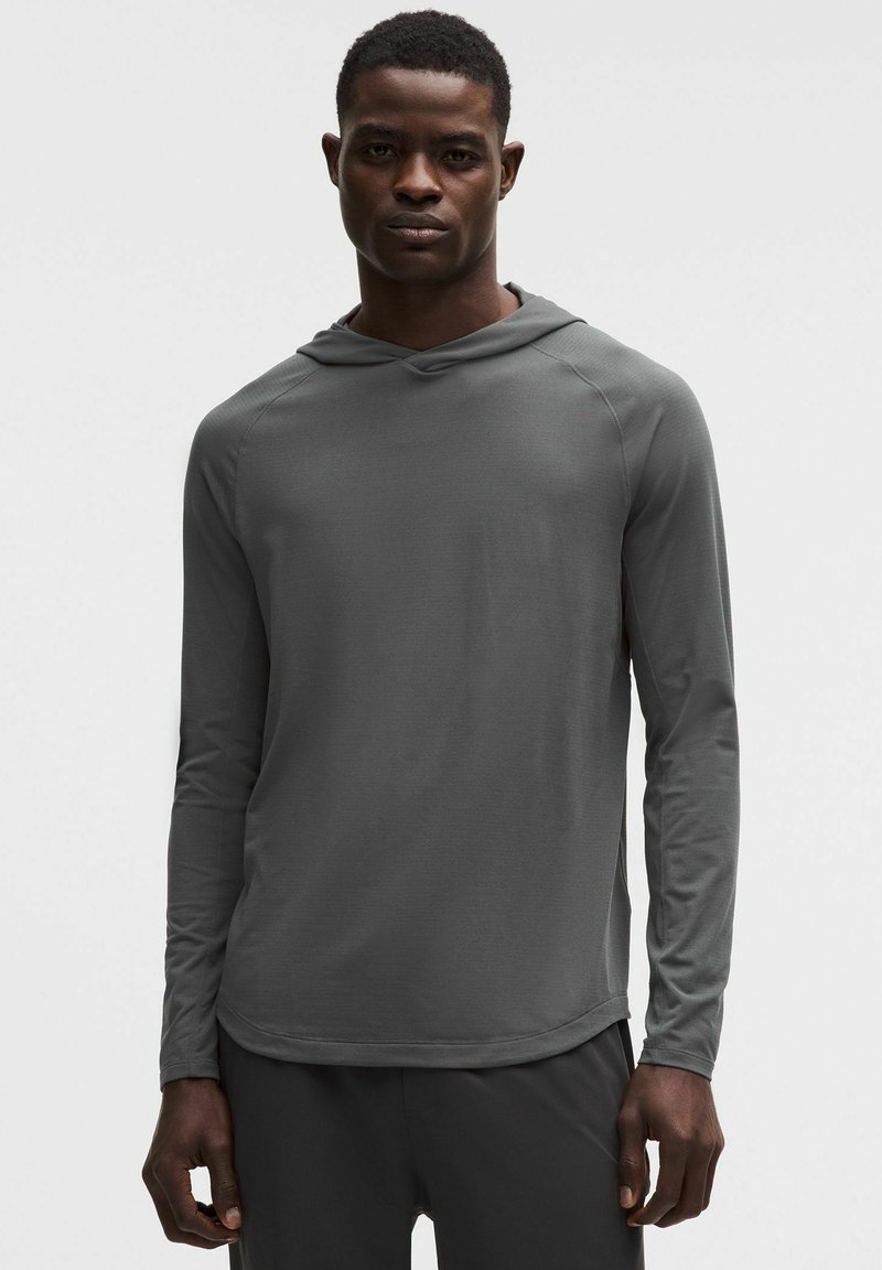 Mężczyzna w ciemnoszarej, długorękawowej koszulce z kapturem i czarnych spodniach stoi na jasnym, gładkim tle, zwrócony przodem z neutralnym wyrazem twarzy.