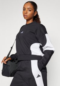 Sweatshirt atlético preto com painéis laterais brancos, apresentando um logótipo da Adidas em roxo. O modelo segura uma mala desportiva preta com uma superfície texturizada.