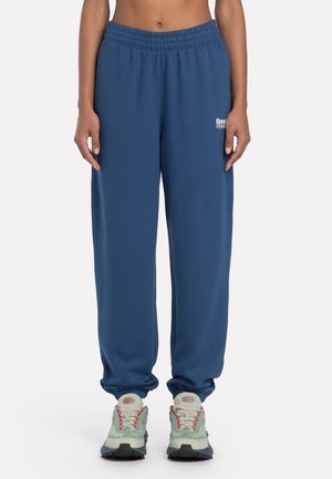 Persoon die blauwe joggerbroeken met elastische tailleband en lichtgroene sneakers met rode accenten draagt, staand tegen een effen achtergrond.