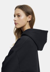 Svart hettegenser med myk tekstur, oversized design, synlige sømmer og kengurulomme. Modellens hår er delvis synlig.