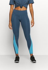 Kvinna stående i blå högmidjade leggings med ljusblå och vita sidopaneler och svarta Nike-sneakers mot en enfärgad bakgrund.