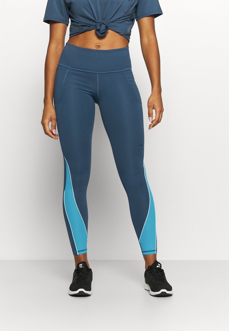 Kvinna stående i blå högmidjade leggings med ljusblå och vita sidopaneler och svarta Nike-sneakers mot en enfärgad bakgrund.