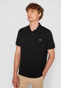 Juodas polo marškinėlis pagamintas iš medvilnės mišinio, turi apykaklę, trumpas rankoves ir mažą siuvinėtą logotipą kairiajame krūtinės pusėje.