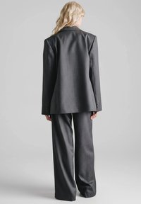 Szary garnitur na miarę, składający się z długiej marynarki z poduszkami na ramionach oraz spodni o szerokich nogawkach. Wykonany z gładkiej tkaniny, prezentujący minimalistyczne detale.