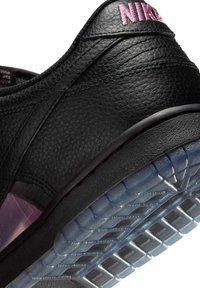 Primer plano del talón de una zapatilla Nike de cuero negro con logo rosa y suela translúcida texturizada sobre fondo blanco.