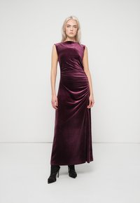 Long burgundy velvet dress with a fitted silhouette, cap sleeves, and draped details on the side, paired with black heeled shoes.