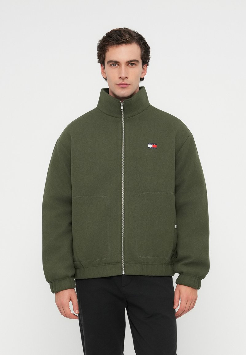 Ελαιοπράσινη ζακέτα με φερμουάρ και ψηλό γιακά, μεγάλες εμπρός τσέπες, ορατές ραφές και μικρή ετικέτα λογότυπο σε κόκκινο, λευκό και μπλε χρώμα.